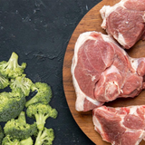 Raw broccoli to the left of raw pork on a wooden chopping board.