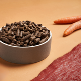 A bowl of DARF Wild Cold Pressed Dog Food beside raw venison meat and raw carrots.