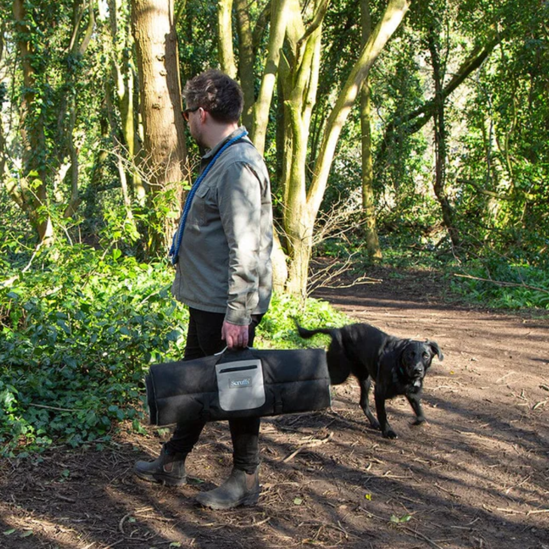 Person carrying a Scruffs Expedition Roll Up Travel Bed, being followed by a black dog in the woods.