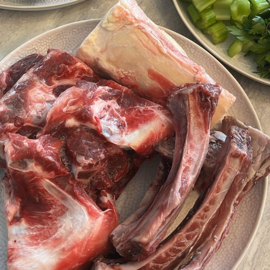 Raw meaty bones for bone broth on a ceramic dish, with greens in the top right hand corner.