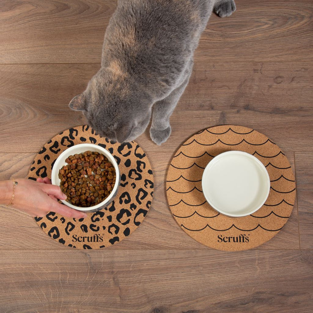 A cat looking into a bowl of food which is sitting on a Scruffs Cork Placemat for pets, with a leopard print design.